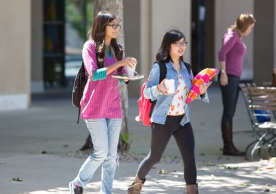 Two female students walk on campus while holding coffees.
