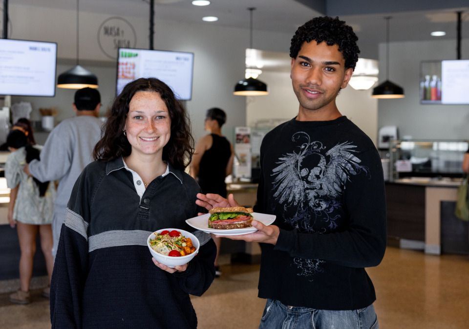 A female and male student wearing black longsleeve shirts hold bowls and plates of food including a salad and sandwich as they stand inside the bistro.