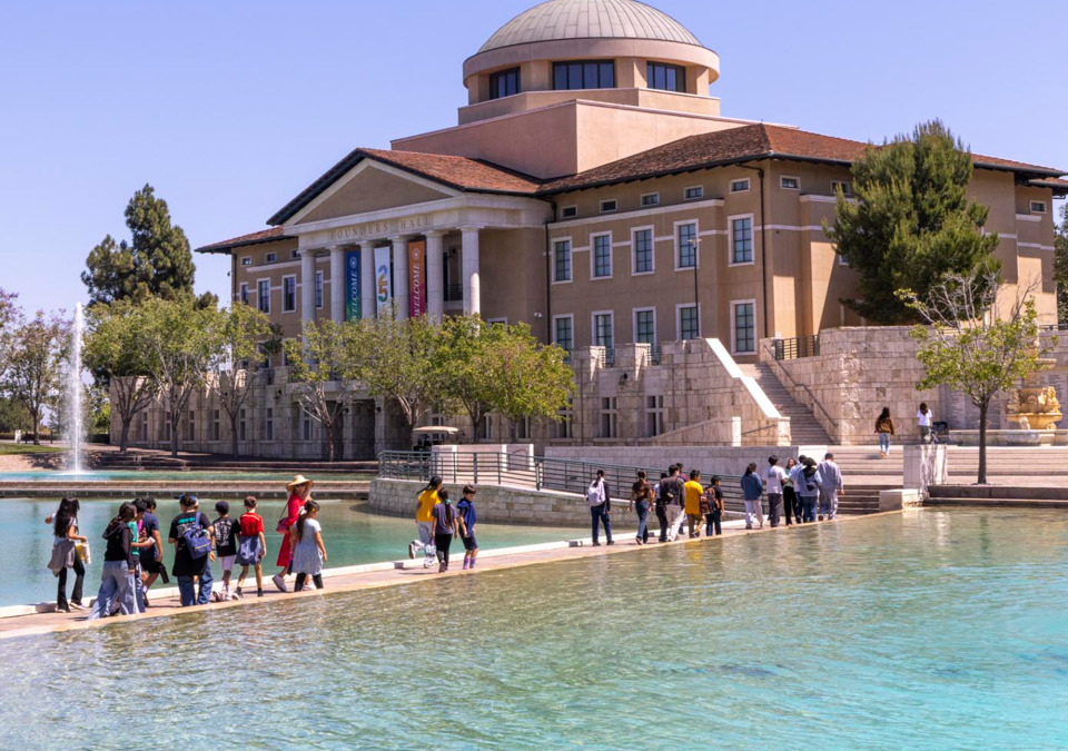A single-file line of students walking across a low walkway over a large, turquoise reflection pool on a sunny campus, heading toward a large, domed building with a fountain visible in the background.