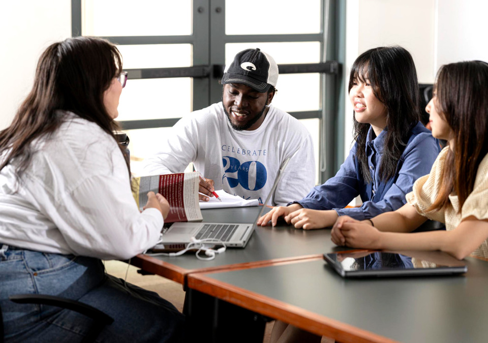 Four students collaborating and talking face-to-face while seated at a classroom table with a laptop and a notebook.