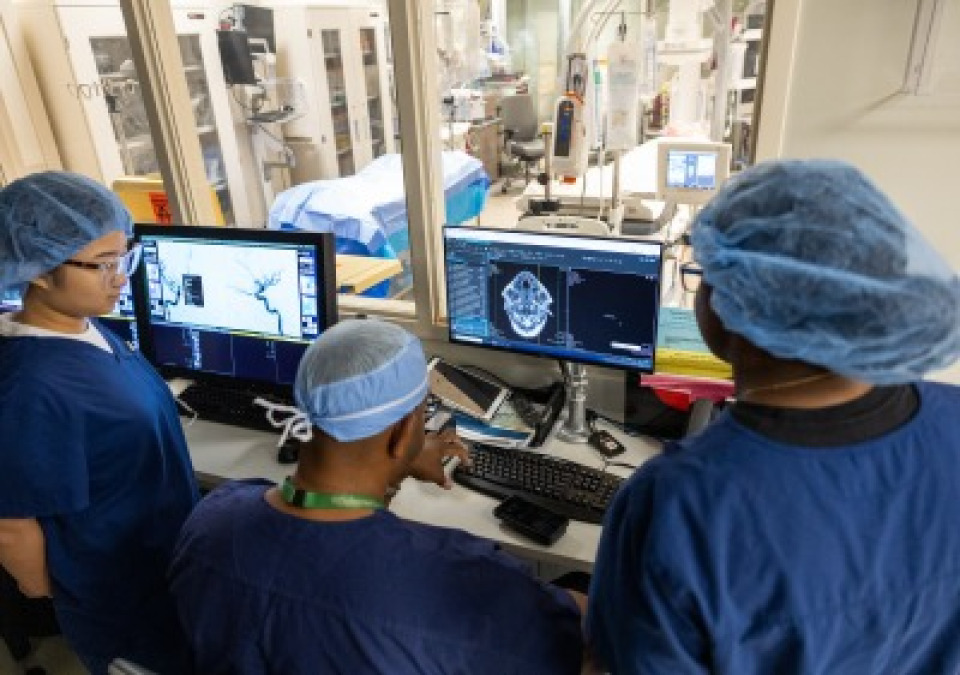 Three medical professionals wearing scrubs and hairnets look at dual monitors displaying medical imaging brain scans. A window behind the monitors looks into a clinical room with medical equipment.