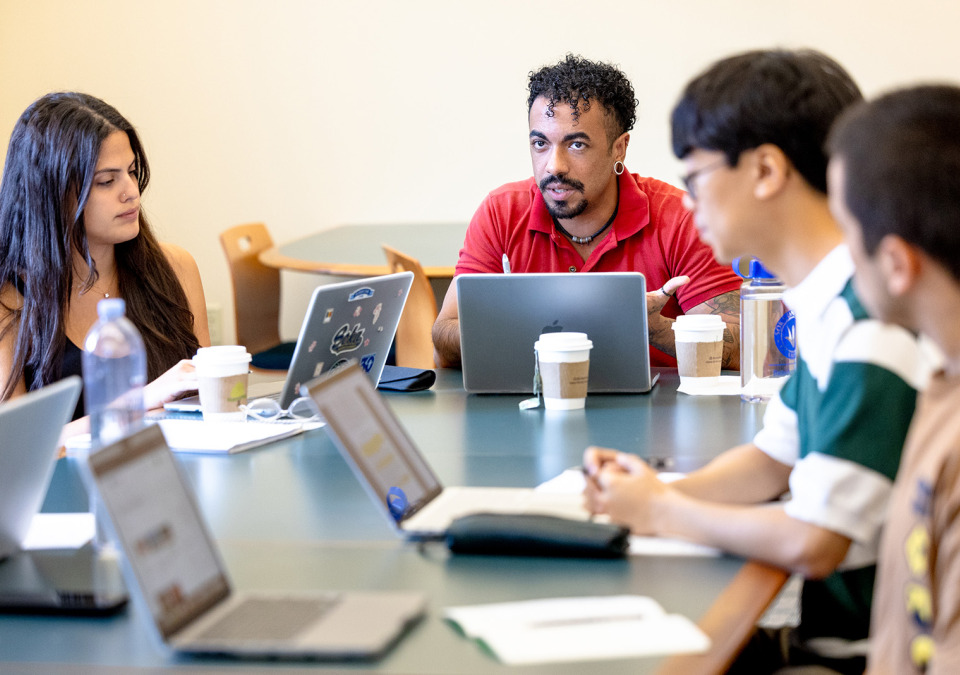 A group of students engaged in a seminar-style discussion around a conference table with laptops and coffee cups.