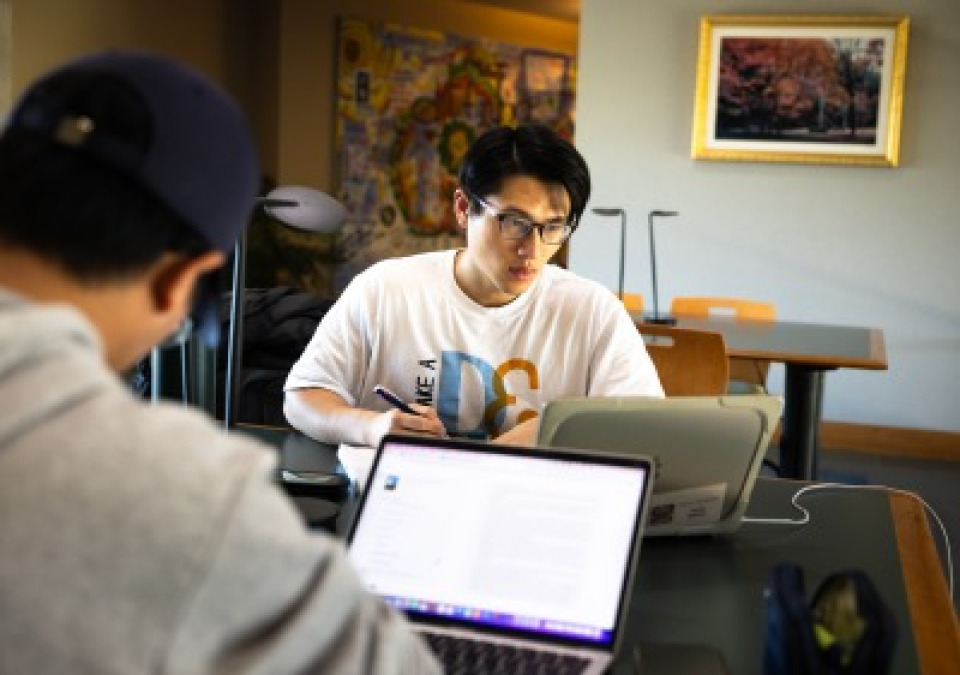 Two students sitting at a long table indoors, focused on their laptops. The student in the foreground wears a dark baseball cap and a grey hoodie, while the student in the background wears glasses and a white graphic T-shirt.