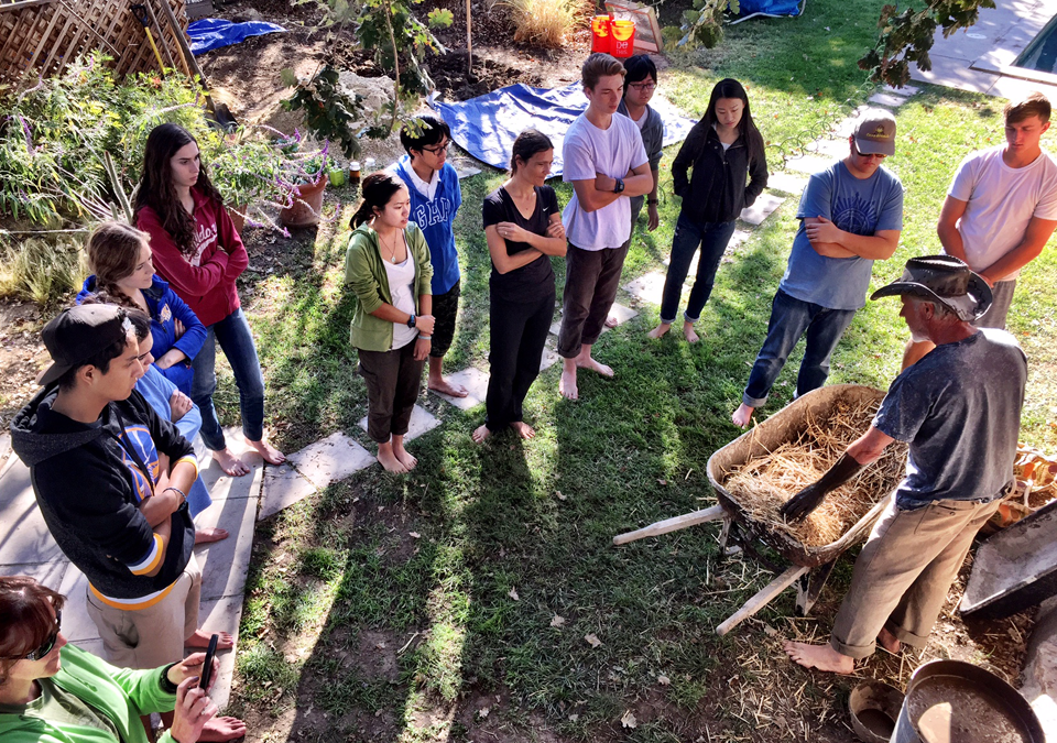 A group of students stand in a grassy yard and listen to an older man with white hear and a cowboy hat as he pulls hay from a wheelbarrow.