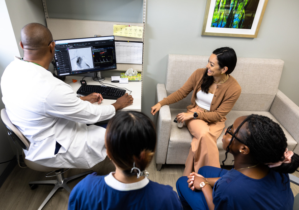 A doctor in a white lab coat shows medical imaging on his computer to two students wearing navy scrubs and an adult female wearing a beige jacket and pants