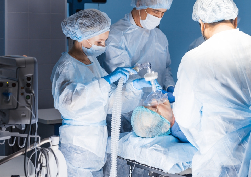Members of a surgical team wearing surgical gowns, caps, and gloves perform surgery. One person is handling the anesthesia equipment.