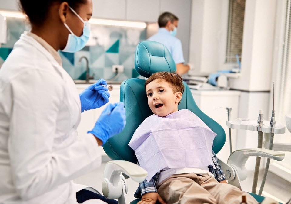 A female dentist wearing a white lab coat and blue gloves holds a dental instrument while a little boy dental patient sits in the chair with a light purple bib over his shirt.