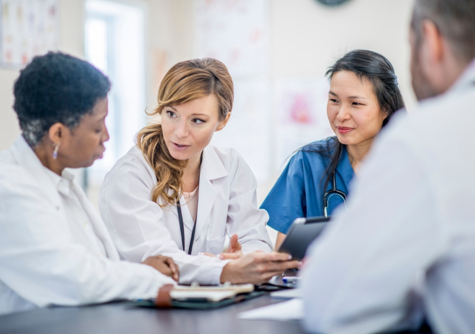 Four medical professionals wearing scrubs, white coats, and stethoscopes have a conversation.