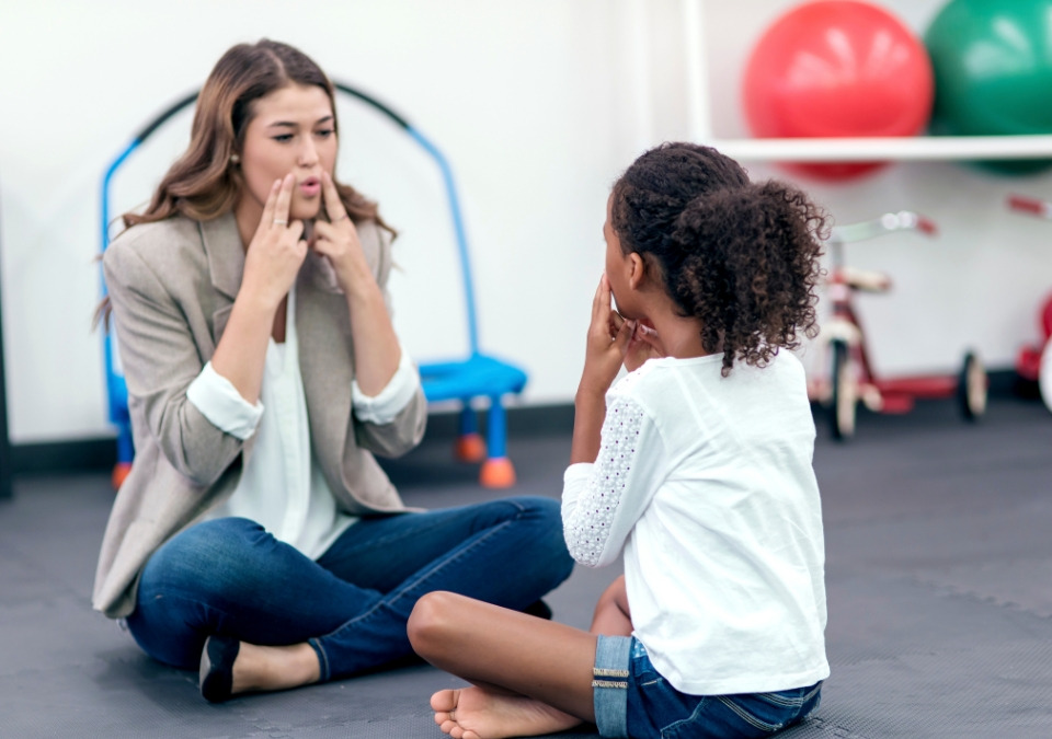 A woman putting her fingers to her cheeks to guide a kid how to spell