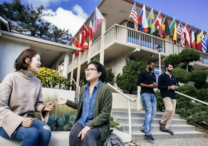 Two female students talk while sitting on a bench beneath a building displaying flags from various countries. Two men are walking down the steps to the side of the women talking.