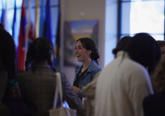A brunette woman wearing a jean jacket smiles as she speaks with other students in front of a variety of international flags.