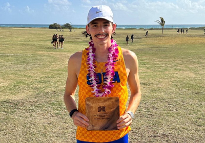 Jordan Bravo is wearing a yellow cross country tank top that says SOKA in navy blue letters with a white baseball hat, and a pink lei around his neck. He is a holding an award.