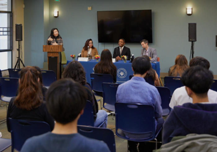 Students attend an event and listen to a woman standing at a podium and a panel of 3 speakers seated at a table to her left.