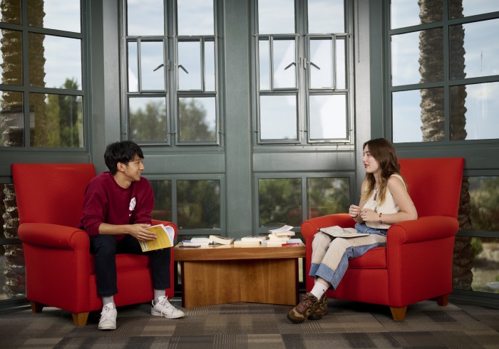 2 students sitting on maroon sofa chairs with a big floor length window and a small coffee table in between them. 