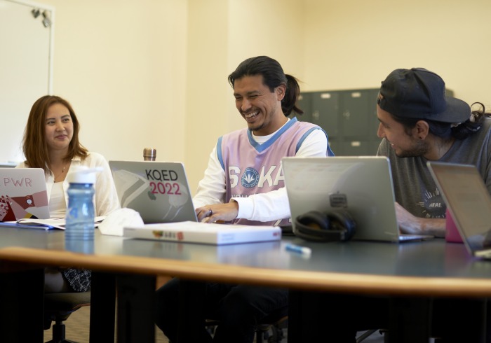 Three graduate students share a laugh as they work on their respective laptops.