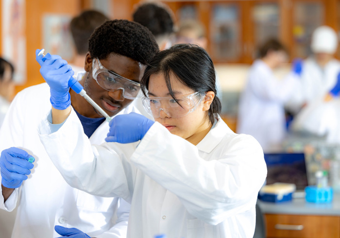 A male and female student in lab coats and safety goggles work together with a micropipette in a science lab.