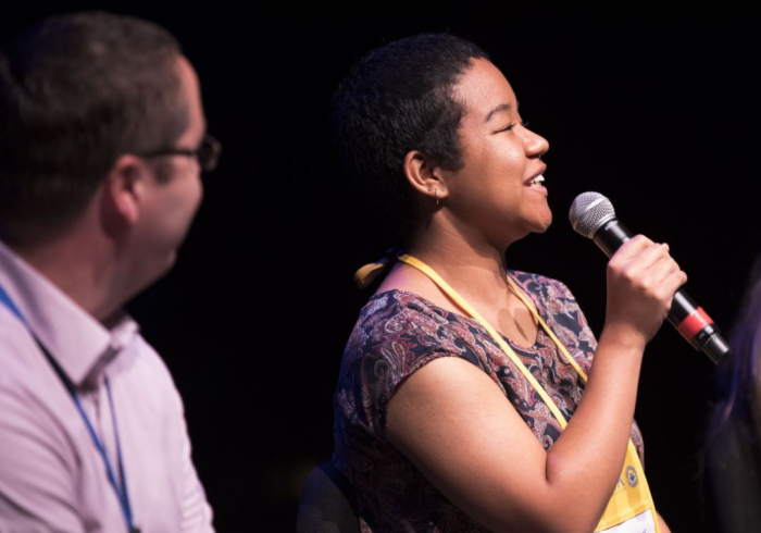 A woman speaks into a microphone while a man listens.