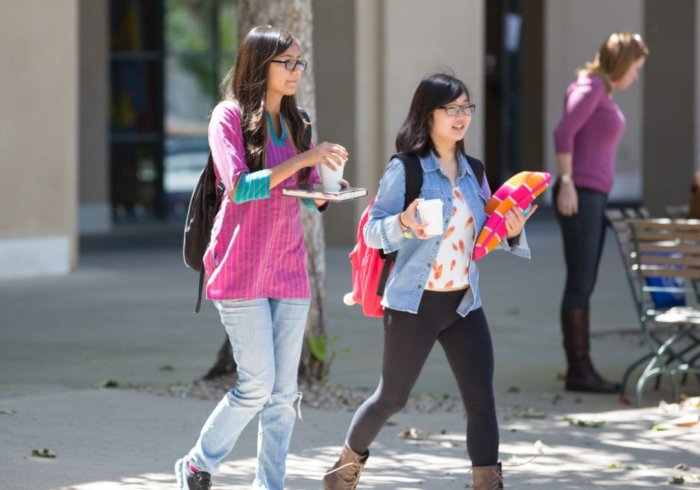 Two female students walk on campus while holding coffees.