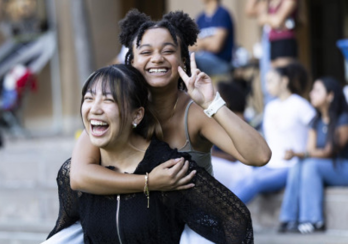 A female student wearing a black lacy zipup and jeans gives a piggyback ride to another female student whose hair is in space buns as she smiles and holds up a peace sign.