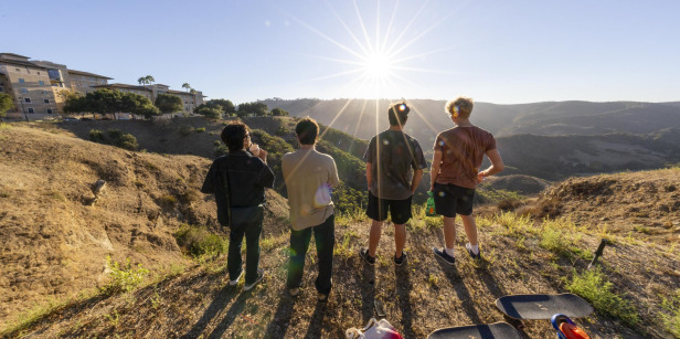 Four male students look out over the canyon while the sun shines brightly on them, the residence halls are to their left.