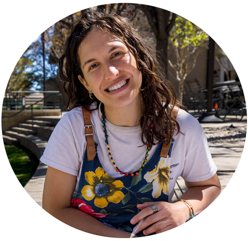 A female student with wavy brown hair wearing a white t-shirt with a floral jumper holds a pen and smiles at the camera as she works outside on SUA's campus.