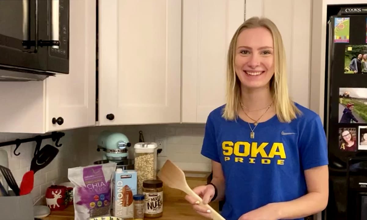 Blond woman wearing blue and yellow Soka T-shirt smiling in kitchen holding wooden spoon