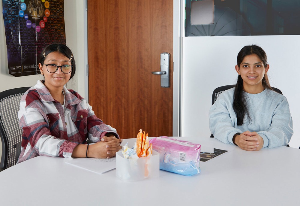 Two women smile at the camera as they sit at a table with menstrual products