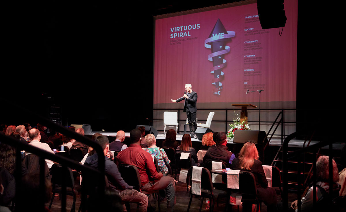 Simon Mainwaring holds a microphone and stands on stage in the Black Box Theatre on Soka's campus where he speaks to an audience about Rewiring Capitalism. There is a screen behind projecting some of his presentation points.