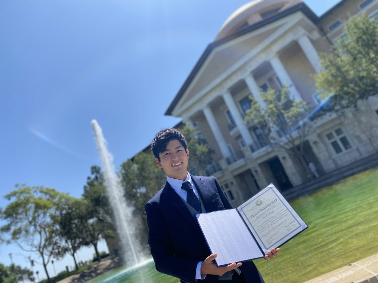 Mark Okuda, winner of the Frederick Douglass Global Fellowship, poses with his Ikeda Scholarship in front of Peace Lake and Founders Hall