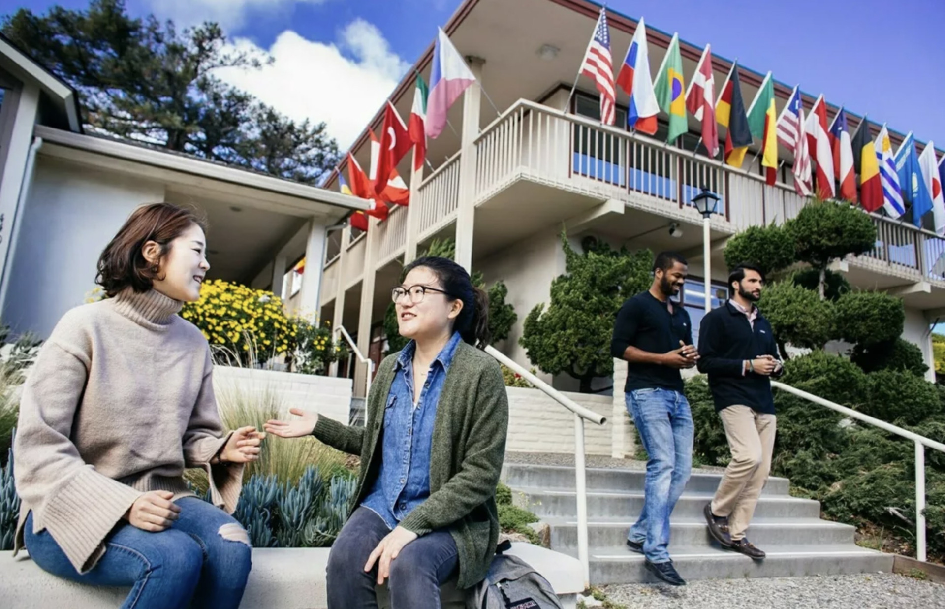 Two female students talk while sitting on a bench beneath a building displaying flags from various countries. Two men are walking down the steps to the side of the women talking.