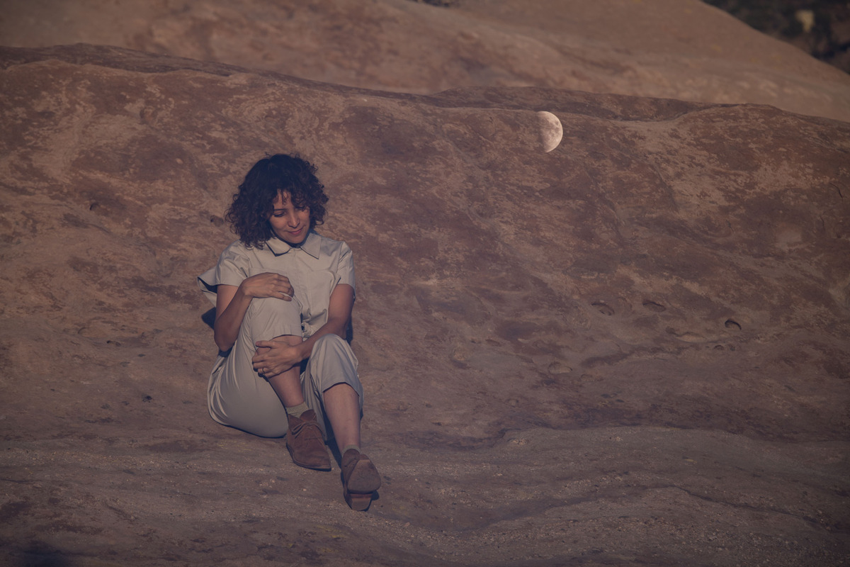 Gaby Moreno sits on a rock surface holding her knee to her chest. A half moon is on the rockface beside her.