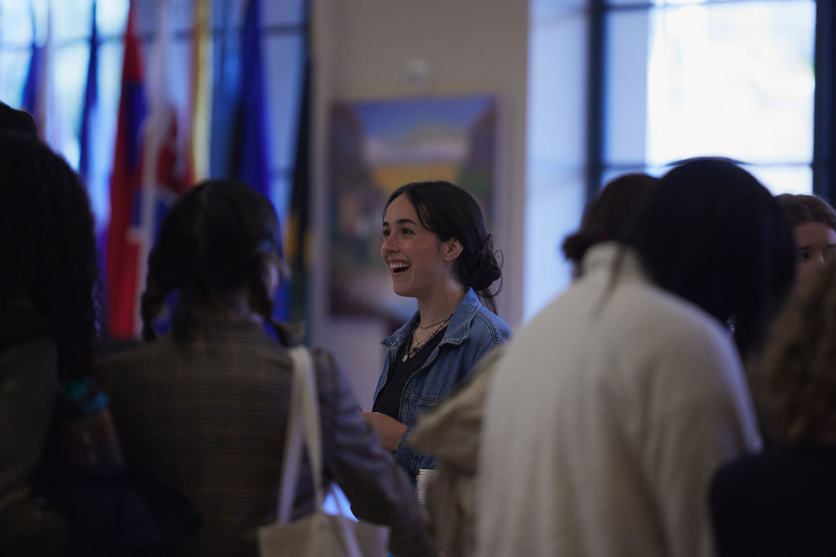 A brunette woman wearing a jean jacket smiles as she speaks with other students in front of a variety of international flags.