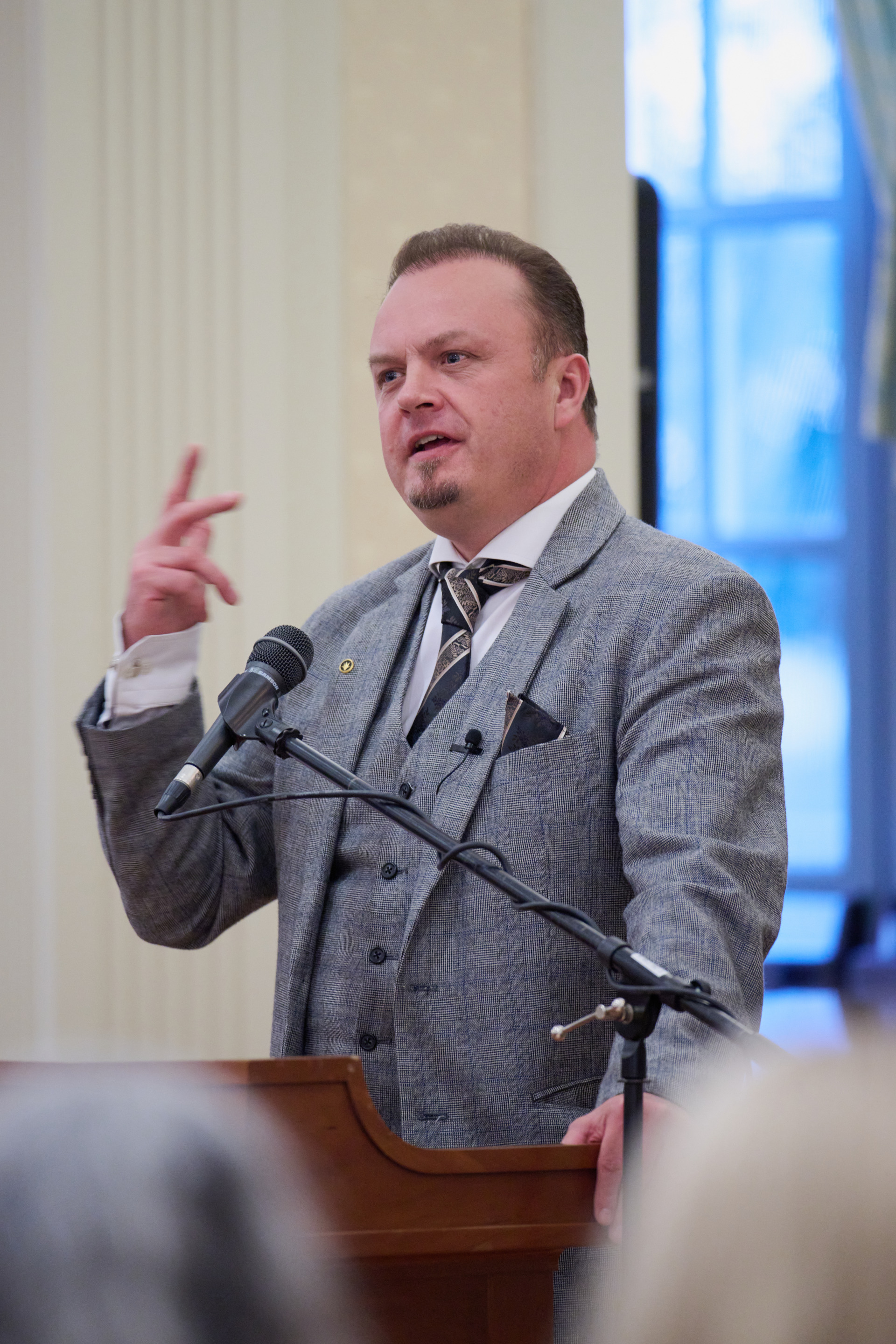 Alexander Harang gestures with his hand as he speaks at a podium with a microphone. He is wearing a grey three piece suit with a black tie.