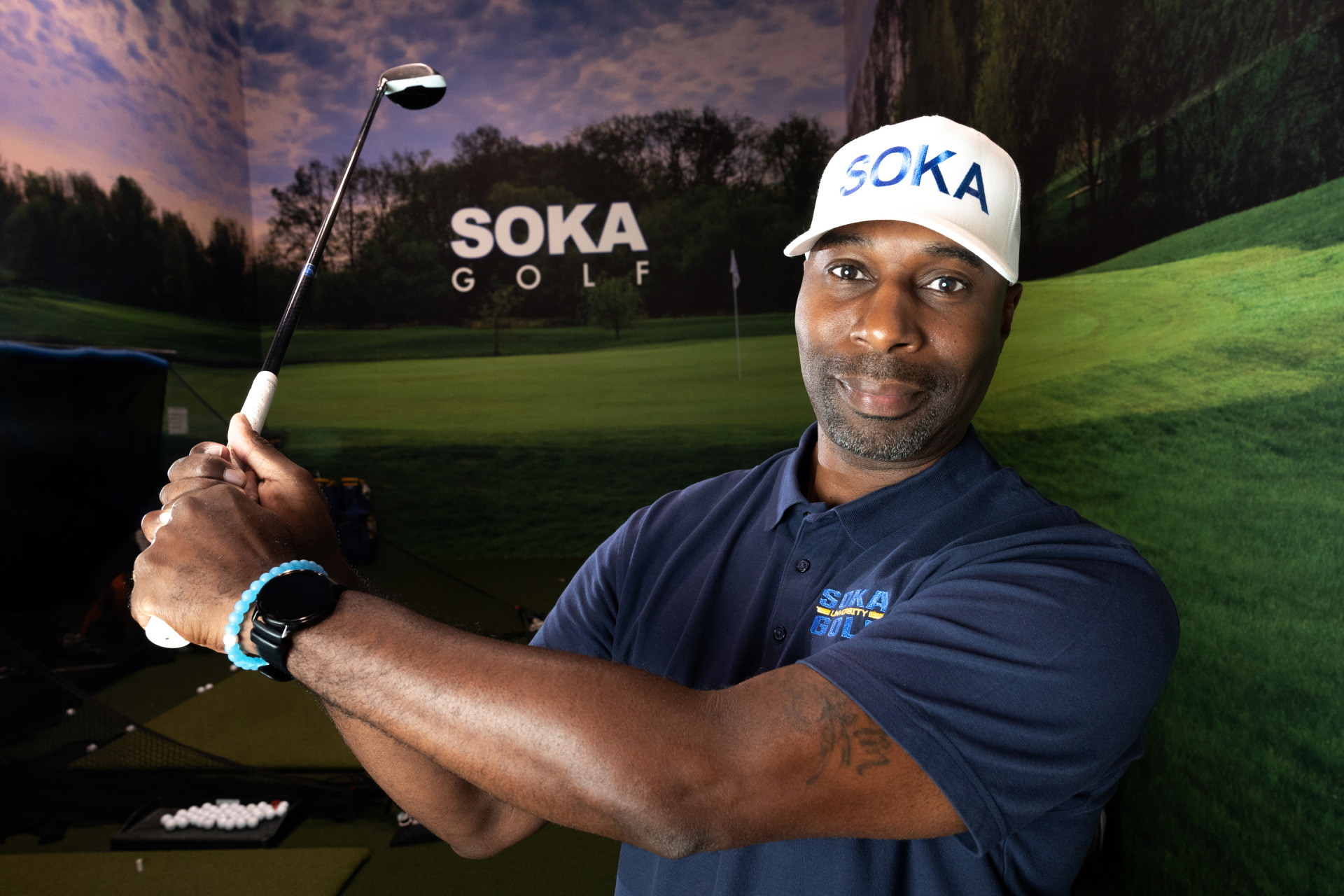 Curtis Robinson, wearing a white Soka baseball hat, holds his golf club and poses in the Soka Golf training room on SUA's campus.