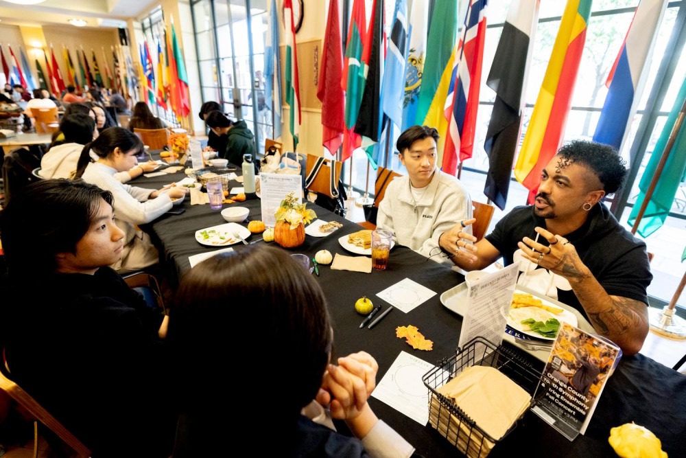 Participants at a "Dialogue Buddies" event engage in conversation around a table, surrounded by international flags and decorated with small pumpkins and flowers.