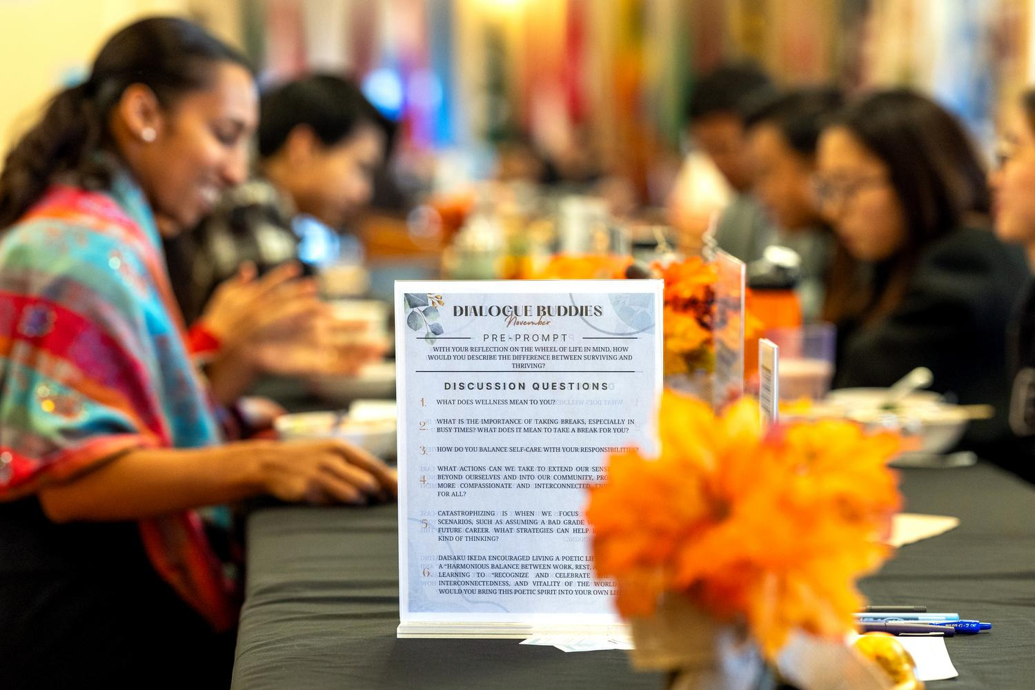 A "Dialogue Buddies" event features reflective questions on wellness and self-care, with participants gathered around a table decorated with orange flowers.