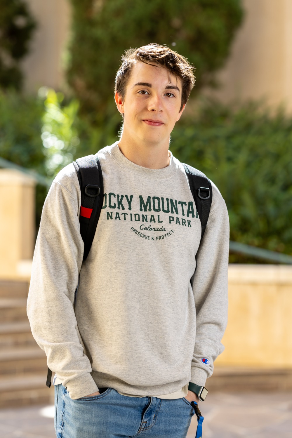 Shea Moloney wears a grey sweatshirt, blue jeans, and backpack and poses for a photo with both hands in his pockets.