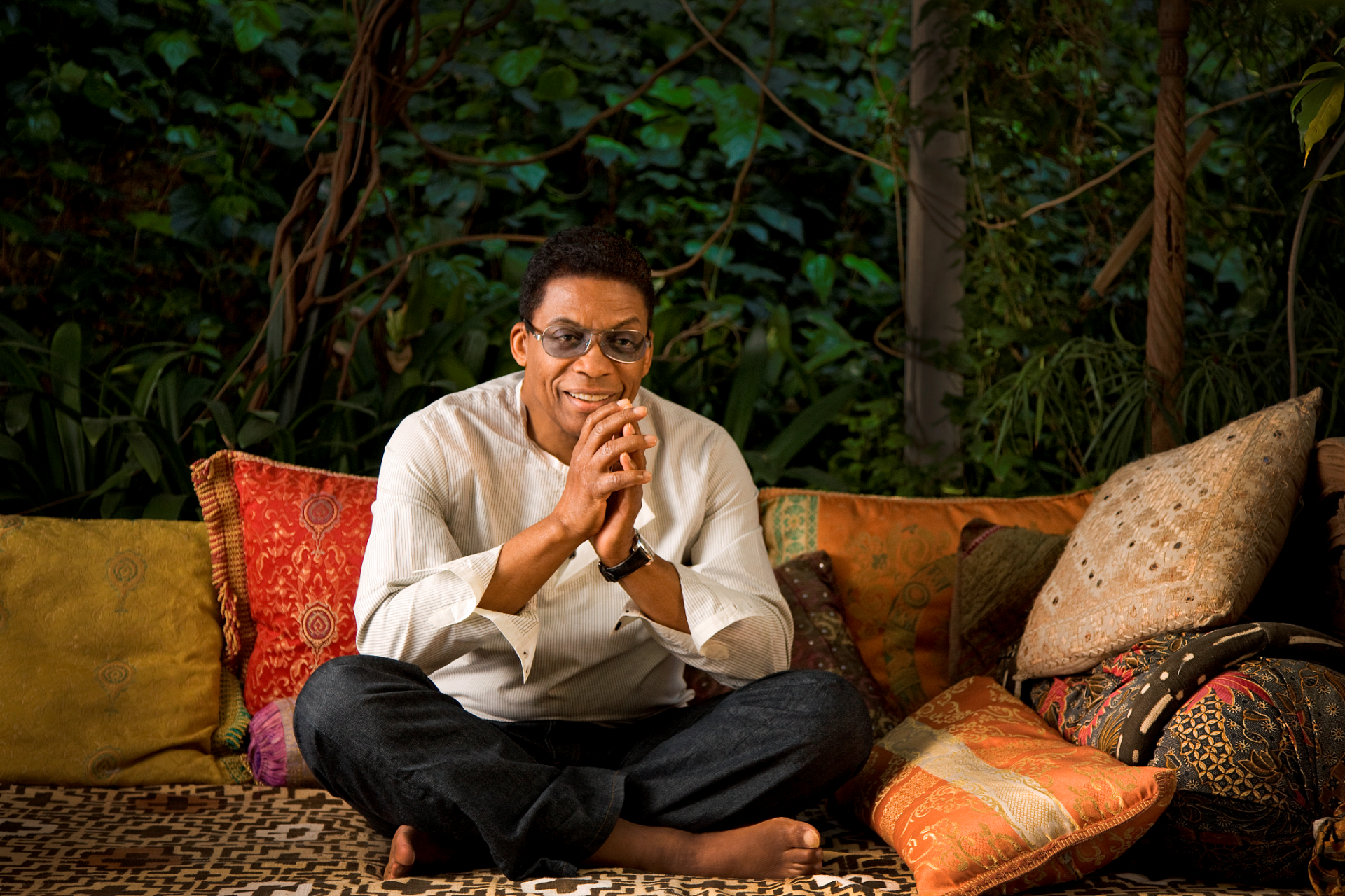 Herbie Hancock sits crosslegged surrounded by colorful throw pillows. He is clasping his hands and smiling at the camera while wearing a white striped shirt, dark jeans, and tinted glasses. Photo by Douglas Kirkland