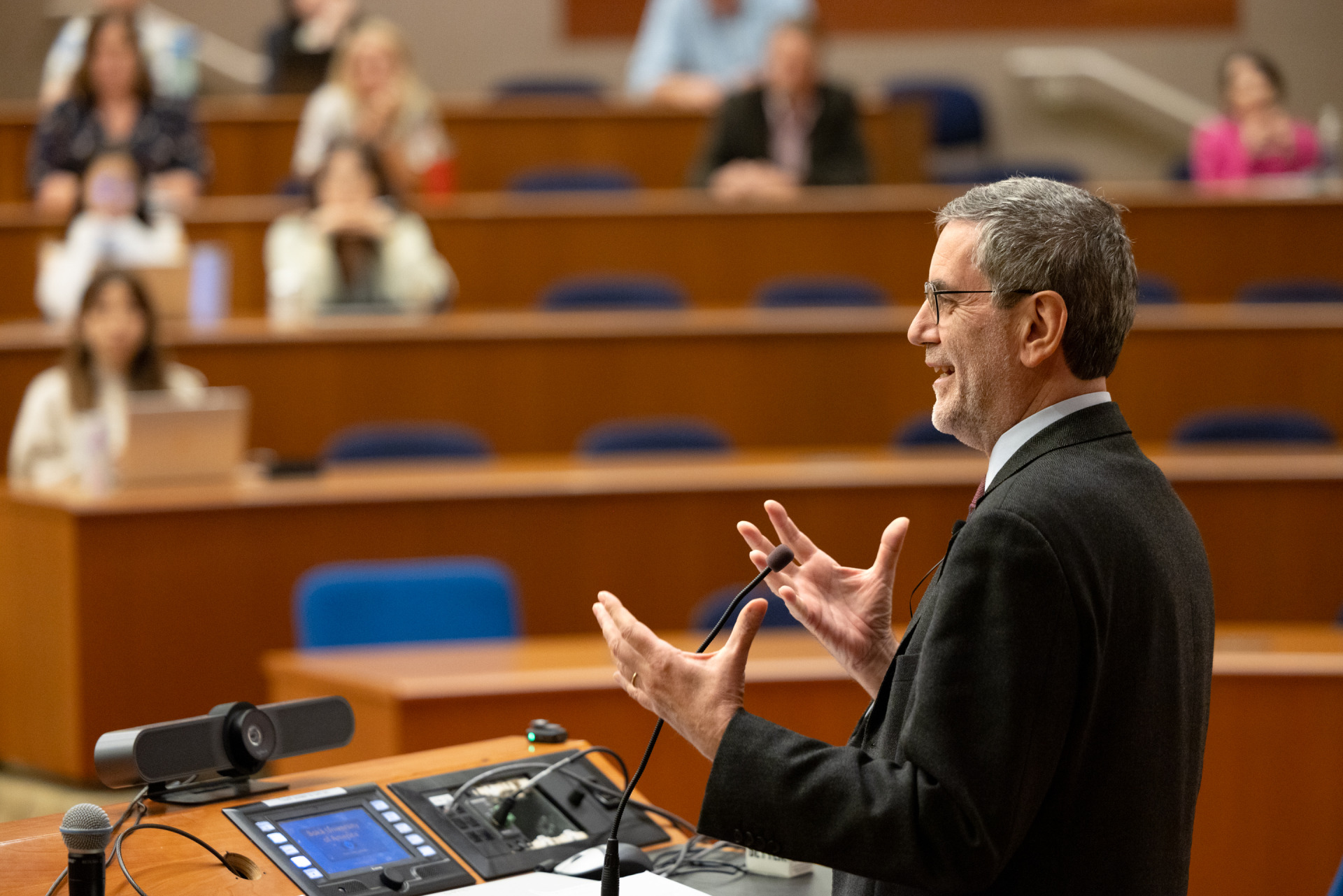 Andrea Bartoli, wearing a suit and glasses, stands at a lectern and gestures with his hands while speaking to an auditorium of people.