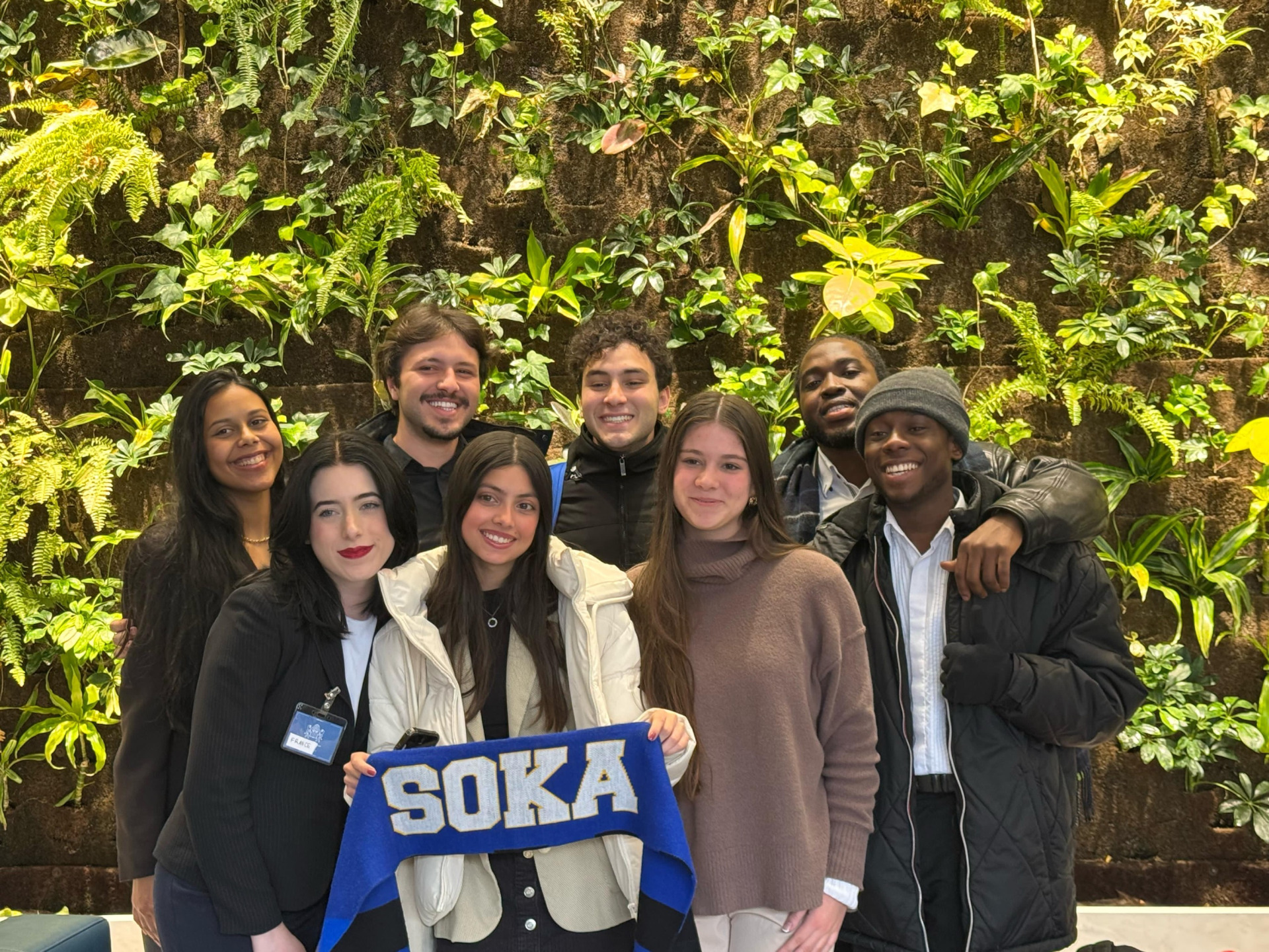 8 student members of the SUA Model U.N. team pose for a group photo in front of a large green wall planter at Toronto's Model U.N. conference. One student is holding up a blue banner that says "SOKA."