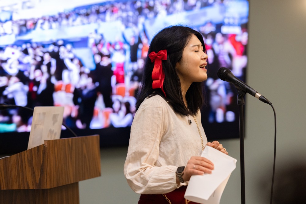 A female student with a red bow in her hair sings into the microphone during the SIGS conference.