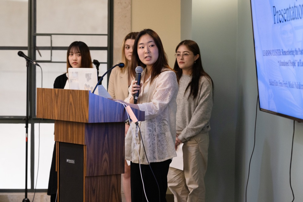 Four female students stand at the podium and give a presentation during a pre-symposium talk.