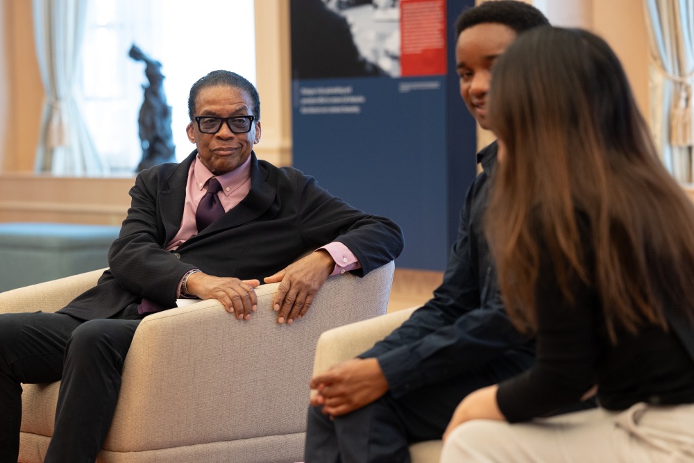 Herbie Hancock sits in a chair and listens attentively to students who are speaking with him.