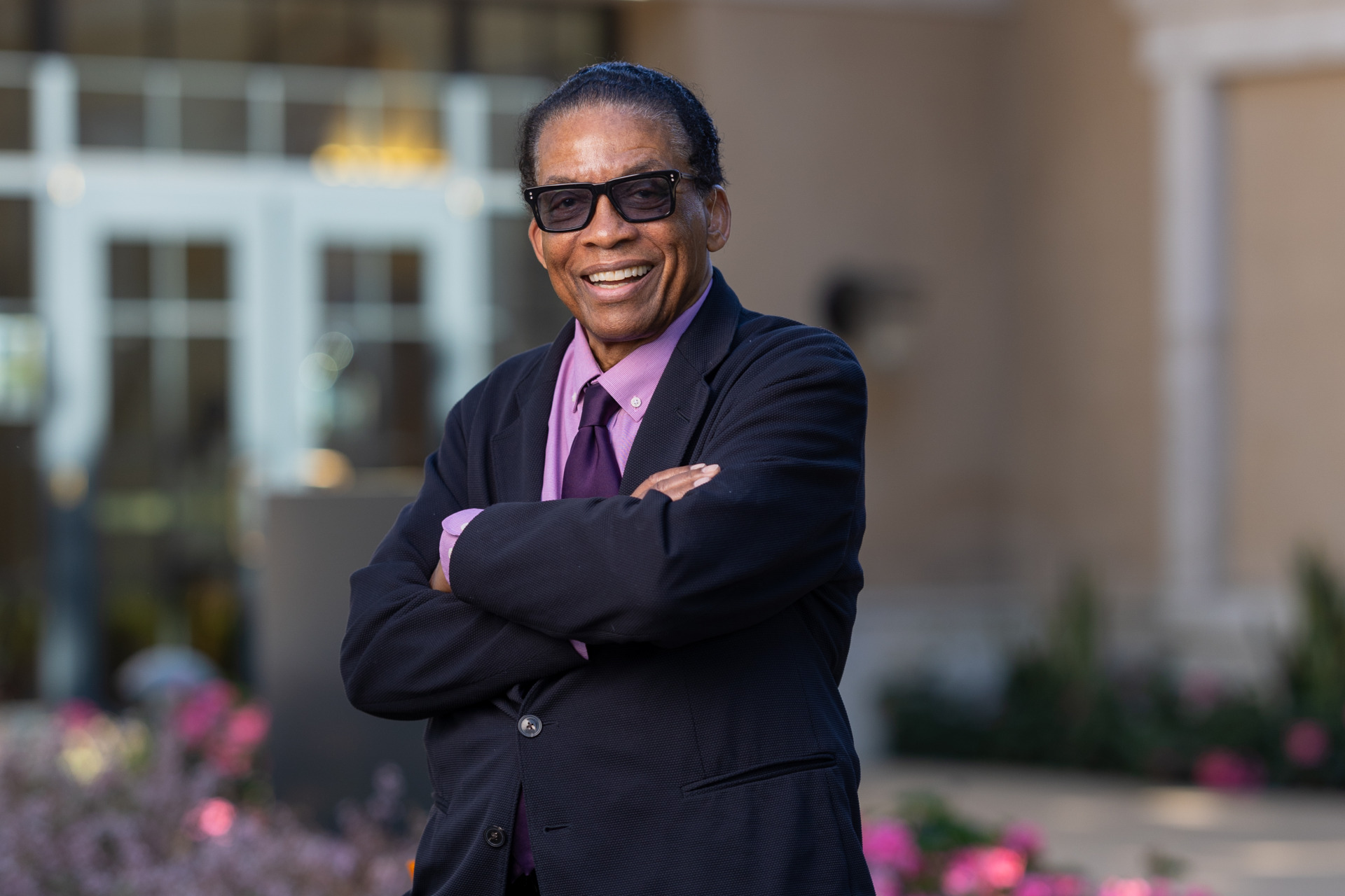Herbie Hancock wears a black suit jacket over a light purple shirt and a dark purple tie. He is wearing tinted sunglasses and smiles as he stands with his arms crossed outside on Soka's campus.