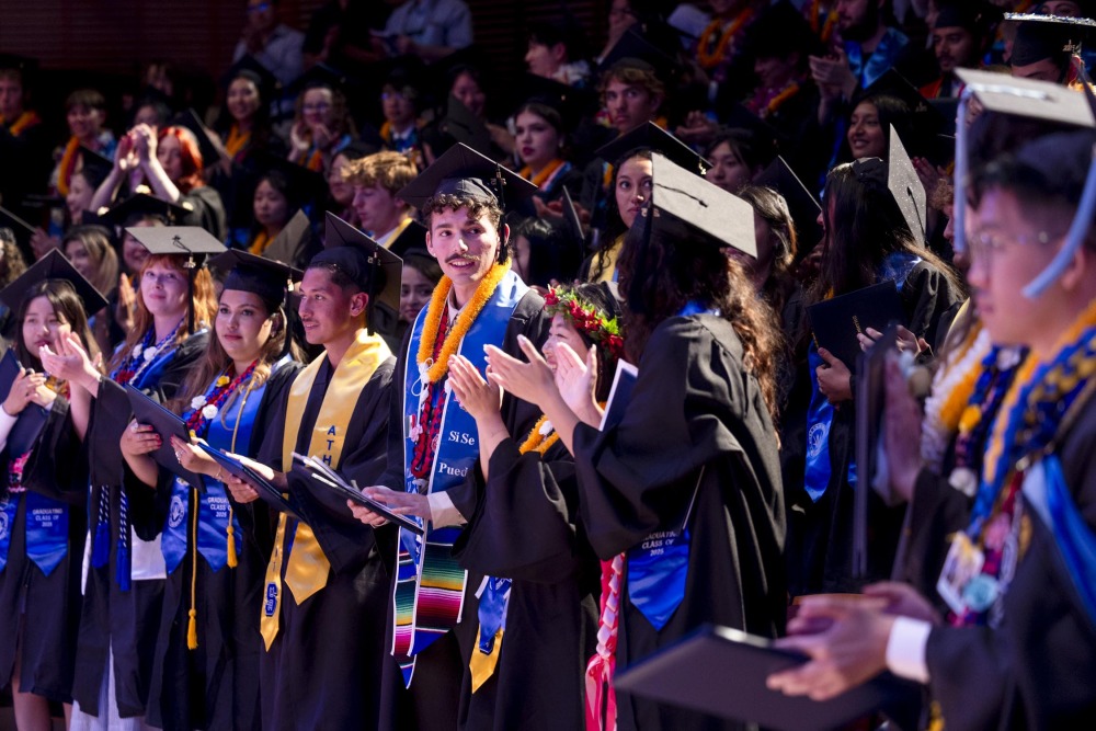 Graduates, wearing their cap and gown, stand and applaud during the 2025 commencement ceremony.