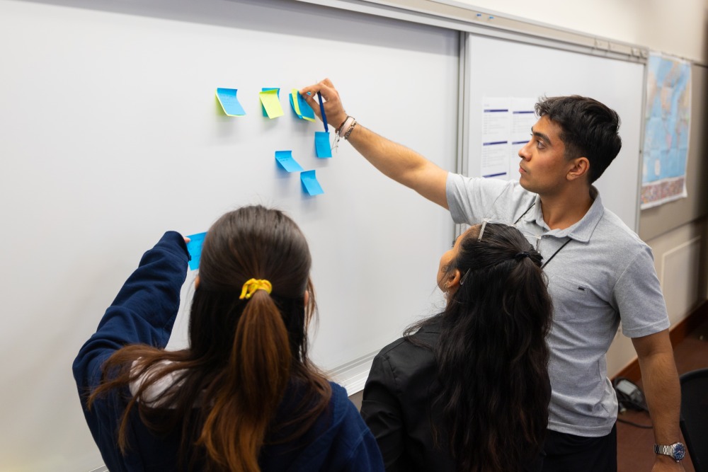 A male student posts a sticky note to a white board while two female students with long brown hair look on.