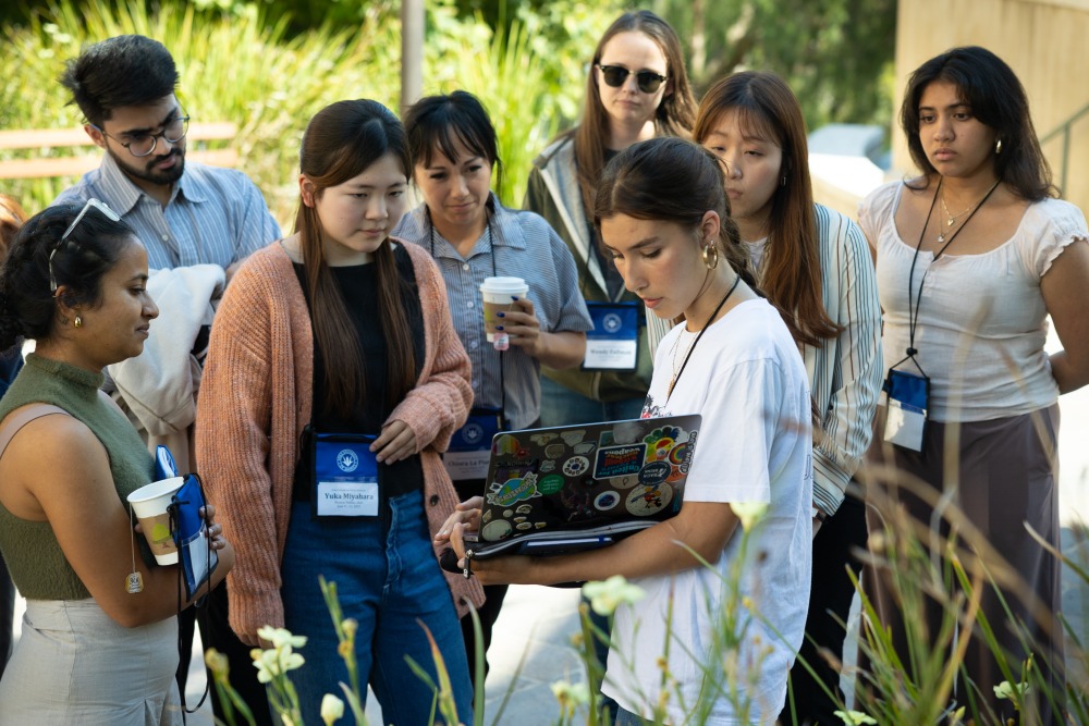 Eight student participants gather around and look at a laptop screen during a break between sessions.
