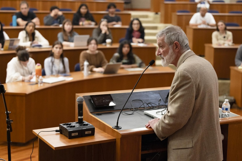 Dr. Ira Helfand wears a tan suit and speaks into a microphone while standing at a lectern and presenting in front of an audience of student participants.