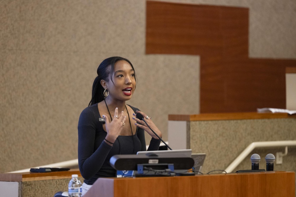A female student speaker wearing a black shirt and red lipstick gestures with her hands while giving a presentation.