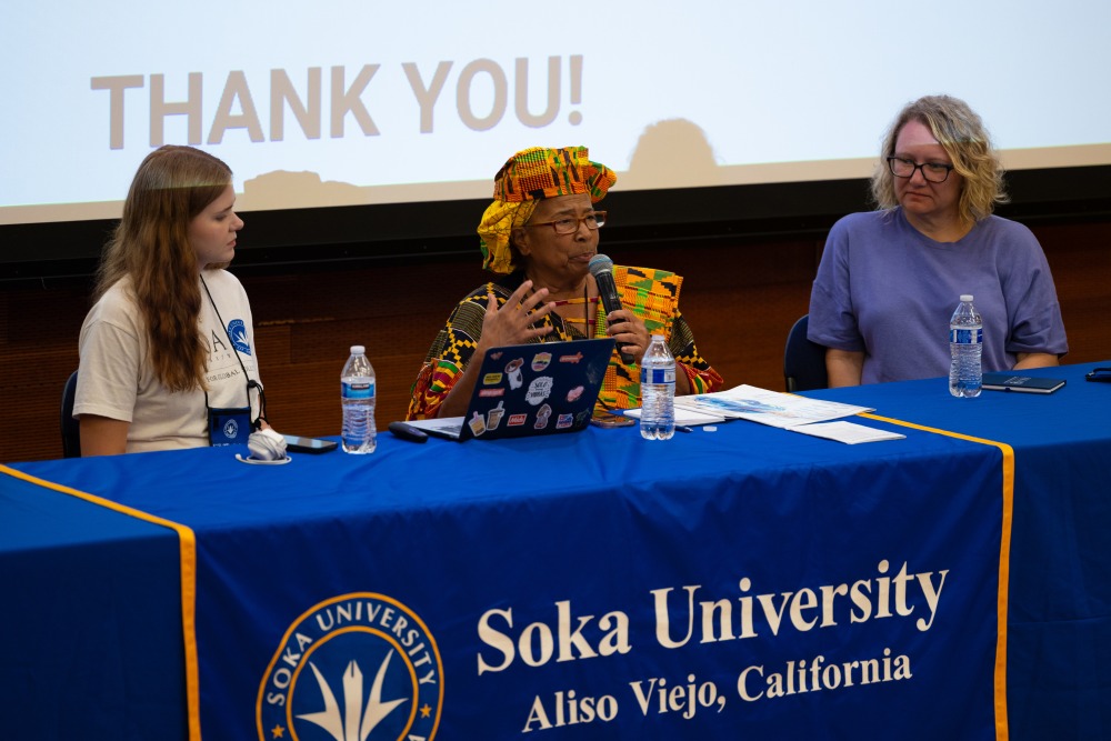Three women sit at a table adorned with a Soka University banner. The woman in the middle speaks into the microphone.